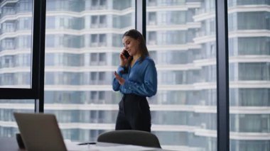 Gülümseyen yönetici cep telefonuyla modern şirket ofisini konuşuyor. Şehir manzaralı panoramik pencereden akıllı telefonu arayan mutlu iş kadını. Güzel kadın çalışanlar arkadaşça telefon konuşmaları yapar..