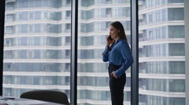 Başarılı bir iş kadını ofisteki panoramik pencerede cep telefonunu arıyor. Şık esmer, şirket stratejilerini cep telefonuyla konuşuyor. Gülümseyen kadın girişimci akıllı telefonla pazarlık yapıyor..
