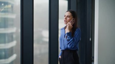 Akıllı telefondan konuşan mutlu girişimci panoramik pencerenin önünden yürüyor. Gülümseyen esmer, ofis binasında cep telefonunu arıyor. Konuşmanın keyfini çıkarın. Güzel iş kadını telefonla iş konuşuyor..
