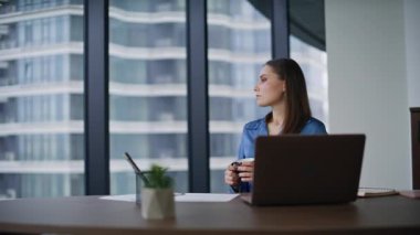Ofis müdürü kahve fincanı tutarak rahatça işyerinde dinleniyor. Panoramik şehir manzaralı sakin bir iş kadını. Şık kadın çalışan, iş yerinde dizüstü bilgisayara bakıyor..
