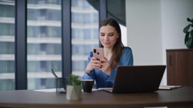 Modern dizüstü bilgisayar işyerinde cep telefonu okuyan gülümseyen bir girişimci. Şirket ofisinde akıllı telefon uygulaması arayan zarif bir esmer. İş yerindeki mutlu iş kadını mesajlaşması..