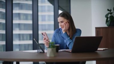 Zarif bir ofis çalışanı cep telefonuna mesaj gönderiyor panoramik pencereli çağdaş bir dolapta oturuyor. Gülümseyen iş kadını akıllı telefon mesajları okuyor. İş yerinde telefon bankacılığı uygulamasında başarılı bir kadın.