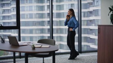 Lüks ofis alanında neşeli, profesyonel bir cep telefonu. Panoramik pencerenin önünde akıllı telefondan konuşan mutlu iş kadını. Gülümseyen esmer şirket işyerinde telefon konuşmasından zevk alır..
