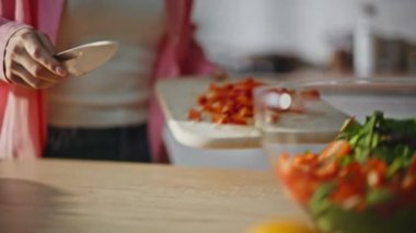 Ahşap mutfak kapısında biber dilimleyen şefin elleri. Kulaklıklı kadın taze kırmızı sebzeleri bıçakla doğruyor ve tabağa koyuyor. Müzik dinleyen kız dairede sağlıklı yemek pişiriyor.