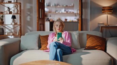 Mutlu bayan evdeki koltukta uzaktan cep telefonu kullanarak internetten alışveriş yapıyor. Haftasonu evde akıllı telefonları dinleyen pozitif sarışın kadın. Genç çekici kız cep telefonu uygulamasından alışveriş yapıyor. 