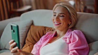 İnternette cep telefonuyla sohbet eden gülen bir kadın modern ışıklı kanepenin önünde yatıyor. Gülümseyen kadın cep telefonuyla video görüşmesinden zevk alıyor. Apartman kanepesinde dinleniyor. Sarışın gevşemiş, akıllı telefon konuşuyor. 
