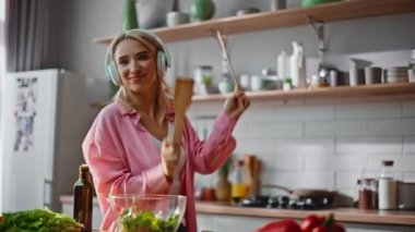Komik kız kulaklıkla yemek hazırlıyor mutfakta en sevdiği şarkı boyunca dans ediyor. Kaygısız neşeli kadın, ahşap spatulalar taşıyan kulaklıkla müzik dinliyor. Gülümseyen sarışın evde salata yapıyor. 