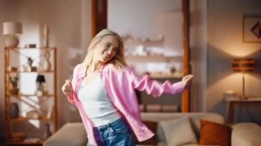 Hafif oturma odasında dans eden, tasasız, tasasız bir kadın. Pembe tişörtlü oyuncu kız evde ritme ayak uyduruyor. Mutlu güzel bayan evde tek başına eğleniyor.