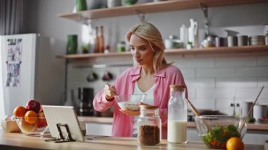 Çevrimiçi kadın kahvaltı tableti yiyor. Mutfağa yakın çekim. Pembe tişörtlü çekici bir bayan tahıl gevreği yiyor ve bilgisayarda film izliyor. Rahat kız hafif mutfakta yemeğin tadını çıkarıyor. 