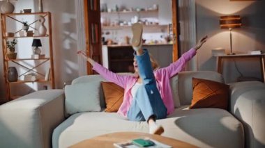 Modern oturma odasındaki yastıklara dokunarak koltuğa düşen memnun kadın. Gülümseyen kız kaygısız bir şekilde hafta sonunun tadını çıkarıyor. Rahat, mutlu bir bayan, koltukta oturmuş, evde tek başına hayal kuruyor.