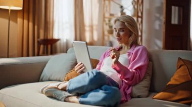 Ev kadını gri koltukta tablet ekrana dokunuyor. İnternetteki bilgisayarını kullanarak apartman dairesinde oturan çekici, gülen bir sarışın. Rahatlamış internet müşterisi alımları uzaktan seçiyor 