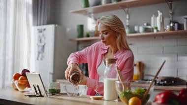 Sabahları mutfakta tablet kullanan bir kadın kahvaltı hazırlıyor. Bilgisayarda video tarifi arayan sarışın kız sağlıklı malzemelerle yemek hazırlıyor. Çağdaş hafif yemeklerde alet izleyen bir bayan.