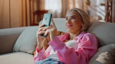 Yumuşak kanepede uzanan ve cep telefonunu izleyen bir kadın. Hayalperest güzel kız dairede sessiz bir hafta sonunda dijital cihazda romantik mesajlar alıyor. Sarışın bayan internetten sipariş vermeyi düşünüyor.