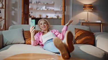 Akıllı telefondan evdeki kanepeye uzanan olumlu kadın videosu. Oturma odasındaki arkadaşıyla uzaktan görüşmede cep telefonu kamerasıyla konuşan gülümseyen kadın. Arkadaş canlısı kız internette sohbet ediyor. 