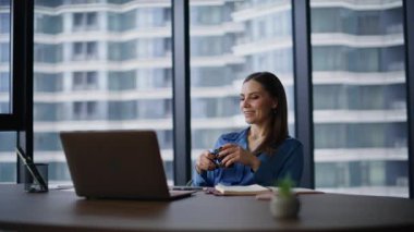 Ofiste oturan zengin bayan elinde kahve fincanı tutuyor. Gülümseyen iş kadını rahat koltuğunda rahatlıyor ve kariyer büyümesini düşünüyor. Kendine güvenen kadın yönetici dizüstü bilgisayarda başarılı olacağını tahmin ediyor..