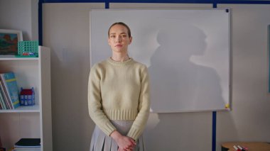 Sınıfta ciddi bir öğretmen kameraya poz veriyor. Portre eğitmeni kadın sakin bir yüz ifadesiyle ayakta duruyor. Kendine güveni tam genç öğretmen doğrudan okul sınıfına bakıyor. 