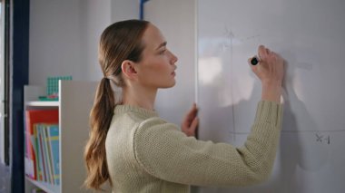 Okul öğretmeni beyaz tahtada matematik problemlerini çözüyor. Işık dolabında çalışan odaklanmış akıllı kadın şemaları çiziyor. Genç eğitimci, tek başına iş yerinde keçeli kalem tutuyor.