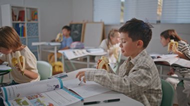 Aç çocuklar için öğle yemeği molası. Yakın çekim. İki çocuk muz yiyor ve masalarda iletişim kuruyor. Sınıfta birlikte boş vakit geçiren komik öğrenciler. Küçük sınıf arkadaşları ders molası veriyor.