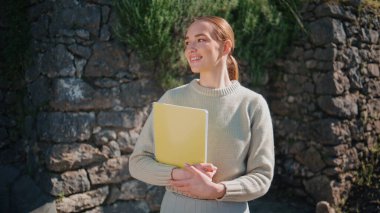 Portre mutlu profesyonel öğretmen. Güneş ışınlarıyla taşlanmış duvarın önünde poz veriyor. Kendine güveni tam, iş belgelerini elinde tutan güzel bir kadın. Kameraya yakından bakıyor. Kitapları olan gülümseyen bayan işçi şehirde dinleniyor. 
