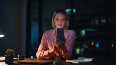 Gece ofisinde gülümseyen bir akıllı telefon. Yakın plan. Zarif bir iş kadını cep telefonunda bankacılık uygulamasına göz atıyor karanlık işyerinde oturuyor. Başarılı bir yönetici maaş telefonu bildirimi alıyor.