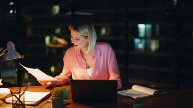 Gece ofisinde lamba ışığında yaratıcı çalışan planlama projesi. Gülümseyen kadın yönetici finansal raporları okuyor ve dizüstü bilgisayar arıyor. Başarılı bir serbest yazar. Uzaktan kumandalı çalışmanın tadını çıkarır. Ofiste kahve içer..