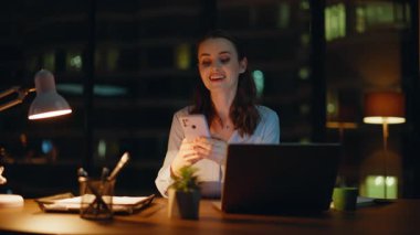 Gece ofisinde akıllı telefondan mesaj atan gülümseyen bir kadın. Karanlık bir iş yerinde dizüstü bilgisayarla cep telefonuna mesaj atan mutlu kadın. Neşeli iş kadını, ara verdiğimizde telefon sosyal medyasında geziniyor..