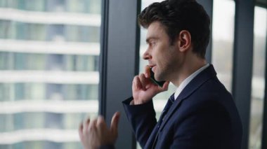 Yakın plan yatırımcı panoramik pencere iş merkezinden cep telefonu görünümlü şehir görüntüsü konuşuyor. Portre işadamı cep telefonuyla projeyi tartışıyor. Yönetmen akıllı telefon konuşmasında görevi açıklıyor