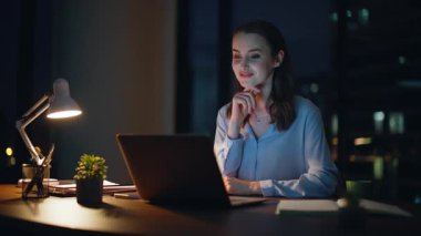 Gece ofisindeki sanal konferansta pozitif çalışan konuşuyor. Gülümseyen iş kadını dizüstü bilgisayardaki web kamerasına el sallayarak video aramasından zevk alıyor. Başarılı yönetici akşam geç saatlerde çevrimiçi toplantıdan memnun.