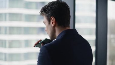 Dalgın yönetici, yüksek katlı ofis penceresinden dışarı bakıyor. Modern kurumsal çalışma alanında finansal stratejileri analiz eden düşünceli bir yönetici portresi. Cep telefonu tutan ciddi bir girişimci..