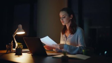 Karanlık dizüstü bilgisayarın iş yerinde başarılı bir girişimci kontrol raporu. Gülümseyen iş kadını bilgisayar ekranı görünümlü dokümanları tatmin etti. Gece geç saatlerde ofiste evrak işleriyle uğraşan kadın yönetici..
