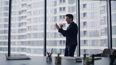 Girişimci cep telefonu haberlerini kutluyor. Yüksek katlı bina panoramik penceresinin yanında duruyor. Heyecanlı iş adamı evet jest yaparak neşeli bir zafer kazanıyor. Çok mutlu bir menajer. Akıllı telefona benziyor. Başarının tadını çıkarıyor..