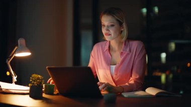 Gece yarısı lamba ışığında çalışan serbest çalışan kadın yakın plan kahve içiyor. Gülümseyen iş kadını karanlık ofiste dizüstü bilgisayara bakan belgeleri kontrol ediyor. Mutlu muhasebeci mali rapordan memnun.