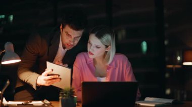 Çalışan, gece ofisindeki kadın yöneticiye tablet verilerini gösteriyor. İki meslektaş akşam bilgisayarın başında mali istatistikleri tartışıyor. E-ticaret projesi üzerinde çalışan ciddi iş adamları.