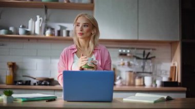 Sabahın erken saatlerinde mutfakta kahve içen yenilenmiş bir kadın. Sarışın kız sıcak bir bardak tutuyor sıcak bir dairede dizüstü bilgisayar izliyor. Günlük kıyafetli bayan yeni bir başlangıcın tadını çıkarıyor.