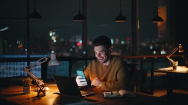 Gülümseyen serbest yazar karanlık dizüstü bilgisayar işyerinde oturan cep telefonu mesajlarını okuyor. Rahat işletme müdürü akıllı telefonda internet iletişiminin tadını çıkarıyor. Gece ofisinde telefon mesajı alan mutlu bir çalışan..