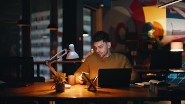 Ciddi programcı lamba ışığı ofisinde oturan dizüstü bilgisayara odaklanmış. Profesyonel serbest çalışan, akıllı telefon görünümlü, gece geç saatlere kadar çalışan. Gece işyerinde traşsız bir iş adamı çalışıyor..