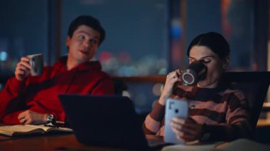 Gece vardiyasında kahve içen gece işçileri yakın plan. Rahatlamış meslektaşlar mesai molasının keyfini çıkarıyorlar. İki sakin iş arkadaşı karanlık iş yerinde dizüstü bilgisayarlarıyla içeceklerini yudumluyor..