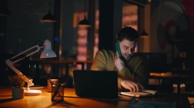 Karanlık ofis alanında cep telefonundan konuşan neşeli bir çalışan. Gülümseyen çevrimiçi yönetici telefon rehberi aracılığıyla sipariş alıyor. Mutlu danışman akıllı telefonu arıyor. Oturma yeri bilgisayarlı iş yeri..