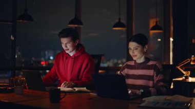 Modern çalışma ofisinde geç saatlere kadar çalışan ekip çalışanları. Gece işyerinde online projede işbirliği yapan iki gülümseyen yönetici. Dost canlısı meslektaşlar dizüstü bilgisayarlarla iş konuşuyorlar..