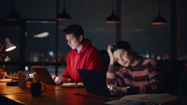 Çok çalışan iş arkadaşları gece iş yerinde dizüstü bilgisayar arıyorlar. Mesaiye kalan iki meslektaşım gece geç saatlere kadar yoğun hissediyorlar. Üzgün ekip çalışanları, karanlık işyerlerinde online iş için motivasyonsuz..