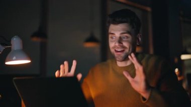 Gece ofisindeki sanal buluşmada gülümseyen serbest çalışan yakın çekim. Bilgisayardaki webcam 'e işaret eden mutlu iş adamı videosu. Yaratıcı adam online konferansta fikirleri tartışıyor.