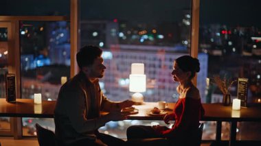 Gece şehir manzaralı restoranda konuşan insanları sevmek. Sıcak kafeteryada romantik bir akşam buluşmasının tadını çıkaran mutlu bir çift. Gülümseyen adam, sevilen kadının elini tutuyor, şefkati hissediyor..