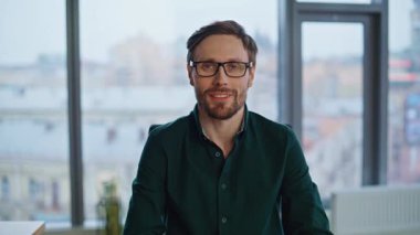 Mutlu yönetici, ofis çalışma alanının yakın planında baş parmağıyla işaret veriyor. Gözlüklü gülümseyen işadamı panoramik pencerenin yanında kameraya bakıyor. Başarılı girişimci şirket işyerinde tek başına oturuyor..