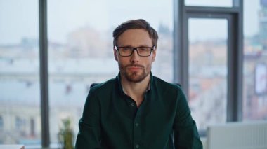 Portre dostu girişimci, modern ofise parmak işareti yapıyor. İş yerinde gözlük takarak poz veren mutlu bir işadamı. Yakışıklı işletme yöneticisi panoramik pencerenin yanında oturuyor.