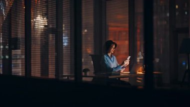 Neşeli iş kadını gece işyerinde akıllı telefon mesajlarına bakıyor. Gülümseyen kadın çalışan cep telefonuyla sosyal medyada sohbet ediyor. Mesai saatleriniz kutlu olsun.. 