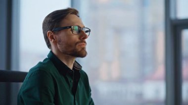 Başarılı yönetici, modern kurumsal iş yerini kamerayla işaret ediyor. Ofis penceresinde poz veren gülümseyen iş adamı portresi. Gözlüklü kendine güvenen yönetici çalışma yerinde oturuyor.