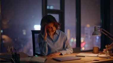 Parlak şehir ışıklarıyla panoramik pencerenin yanında karanlık bir ofiste oturmaktan yorgun düşmüş bir yönetici. Yorgun iş kadını çalışma masasında şakaklara dokunuyor. Şirket profesyoneli işini bitir. Kapalı alanda bitkin hisset..