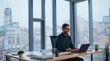 Ofis uzmanı panoramik pencereli çağdaş iş yerinde online projeden memnun. Gülümseyen iş adamı dizüstü bilgisayarın tadını çıkarıyor. Yaratıcı girişimci çalışma alanında başarılı hissediyor.
