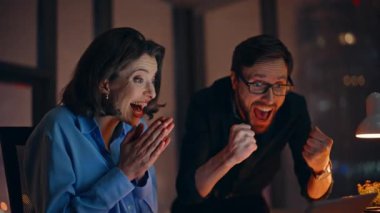 Karanlık iş yerinde dizüstü bilgisayarı görünce sevinen iş arkadaşları. Gülümseyen iş arkadaşları duygusal olarak harika haberlere tepki veriyorlar. Heyecanlı ortaklar beş başarılı takım çalışması veriyor.