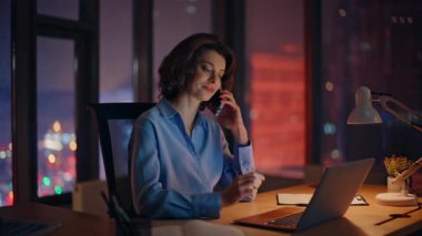 İş kadını gece ofisindeki cep telefonunu arıyor. Kapatma. Gülümseyen iş kadını karanlık işyerindeki akıllı telefondan konuşan dizüstü bilgisayarlı veriler. Pozitif kadın yönetici telefonda fazla mesai yapıyor.